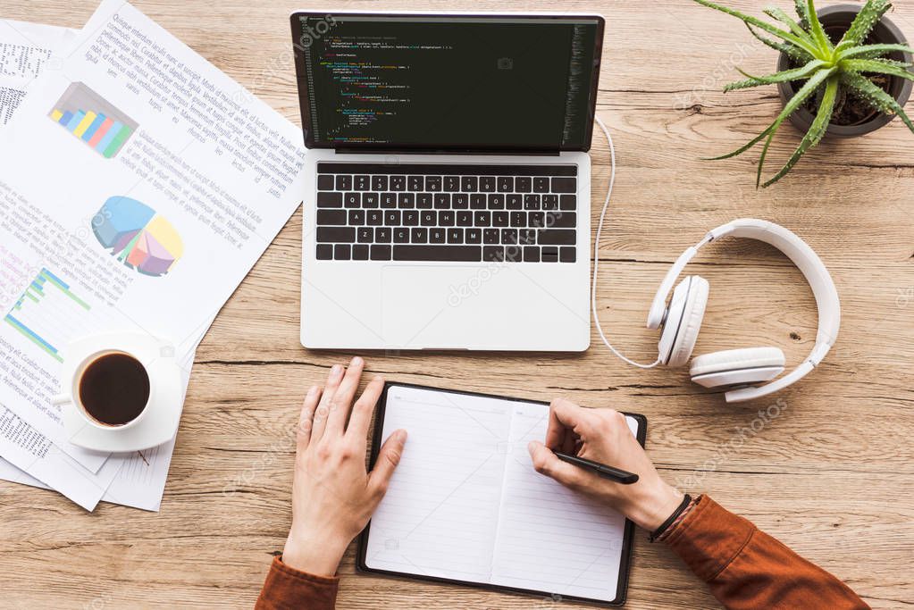 Partial view of man making notes in notebook at workplace with laptop, papers, cup of coffee and headphones