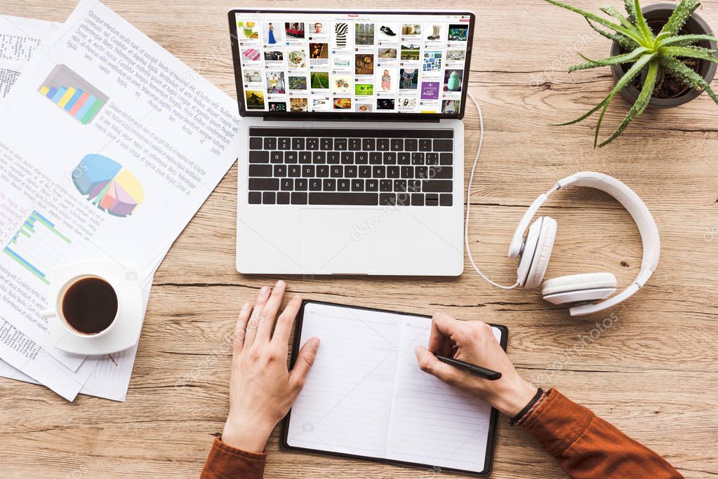 Partial view of man making notes in notebook at workplace with laptop with pinterest website, papers, cup of coffee and headphones
