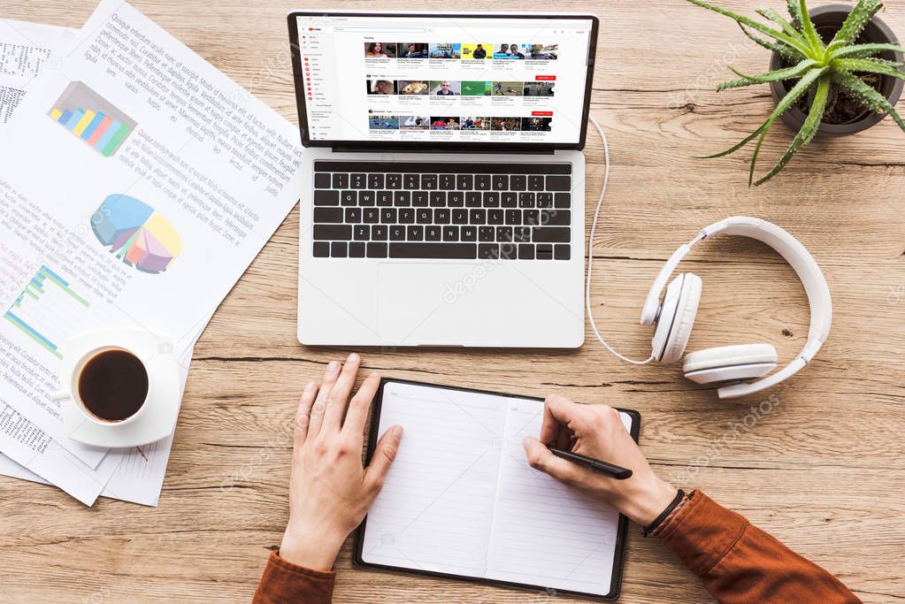 Partial view of man making notes in notebook at workplace with laptop with youtube website, papers, cup of coffee and headphones