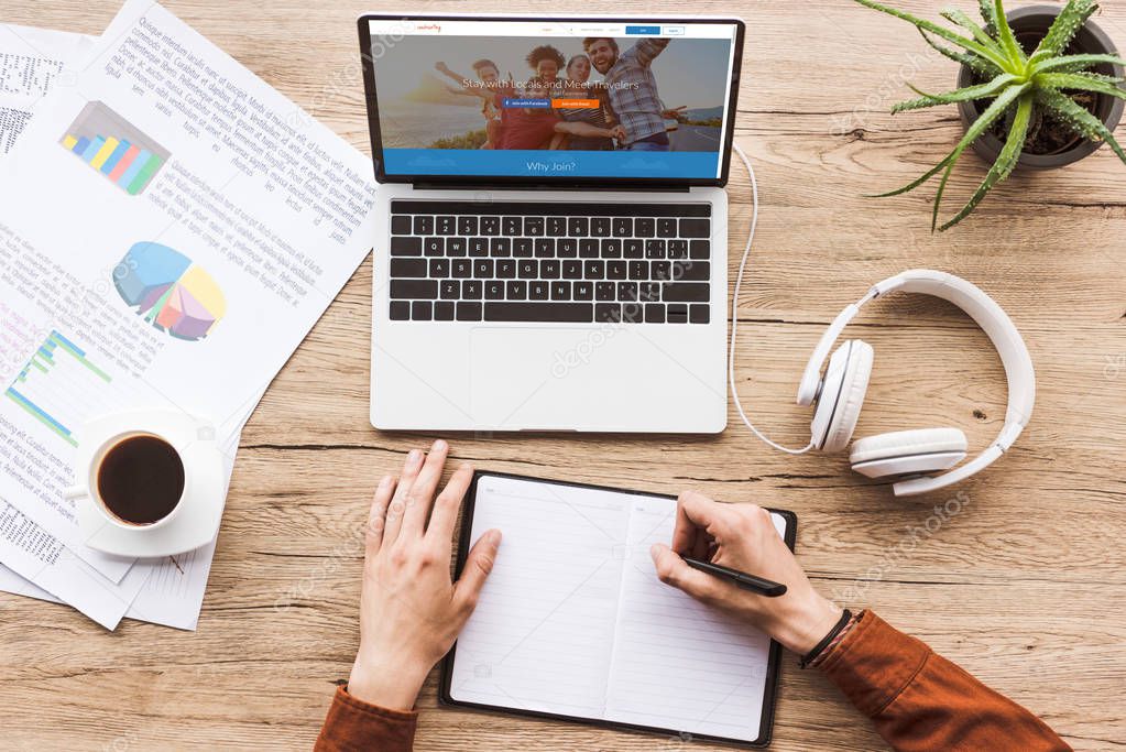 Partial view of man making notes in notebook at workplace with laptop with couchsurfing logo, papers, cup of coffee and headphones