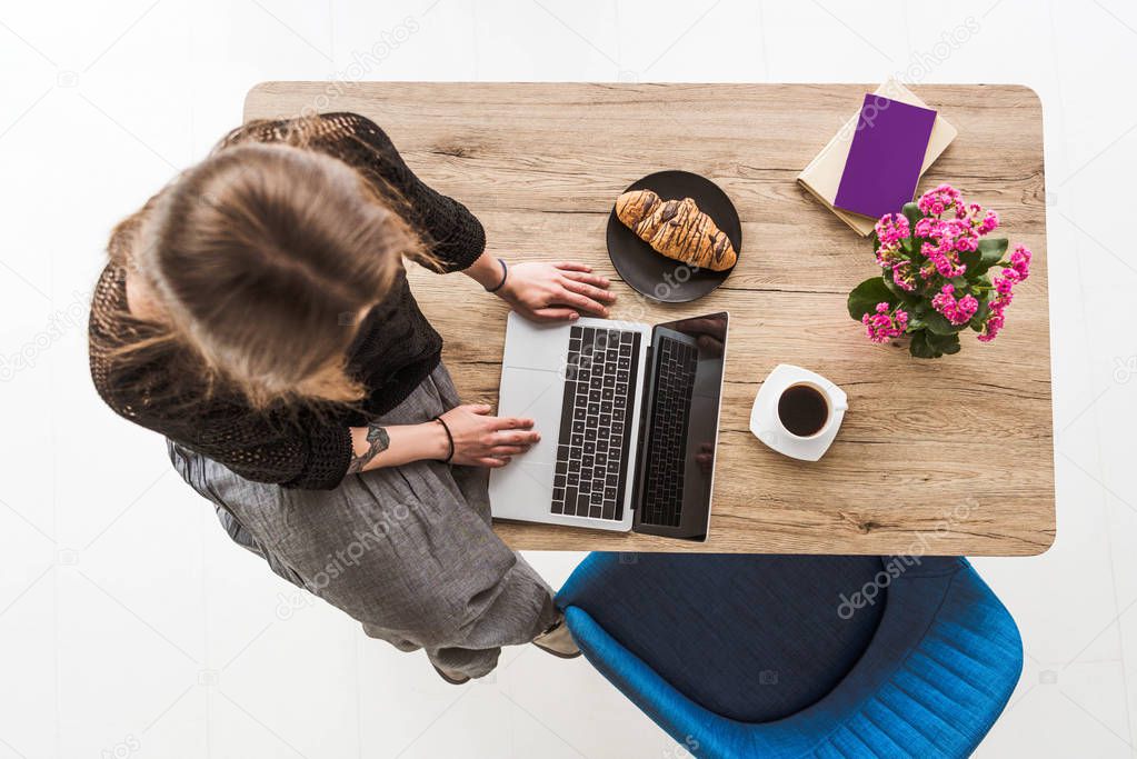 overhead view of woman typing on laptop with blank screen at table with croissant, coffee, flowers, book and textbook 