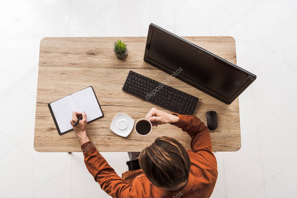 top view of man drinking coffee and writing in textbook at table with potted plant, computer, computer keyboard and computer mouse