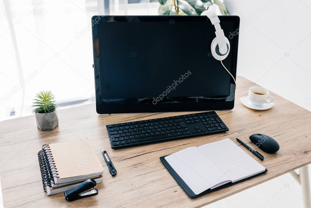 closeup view of headphones on computer monitor, empty textbook, stationery knife, stapler, coffee cup and potted plant 