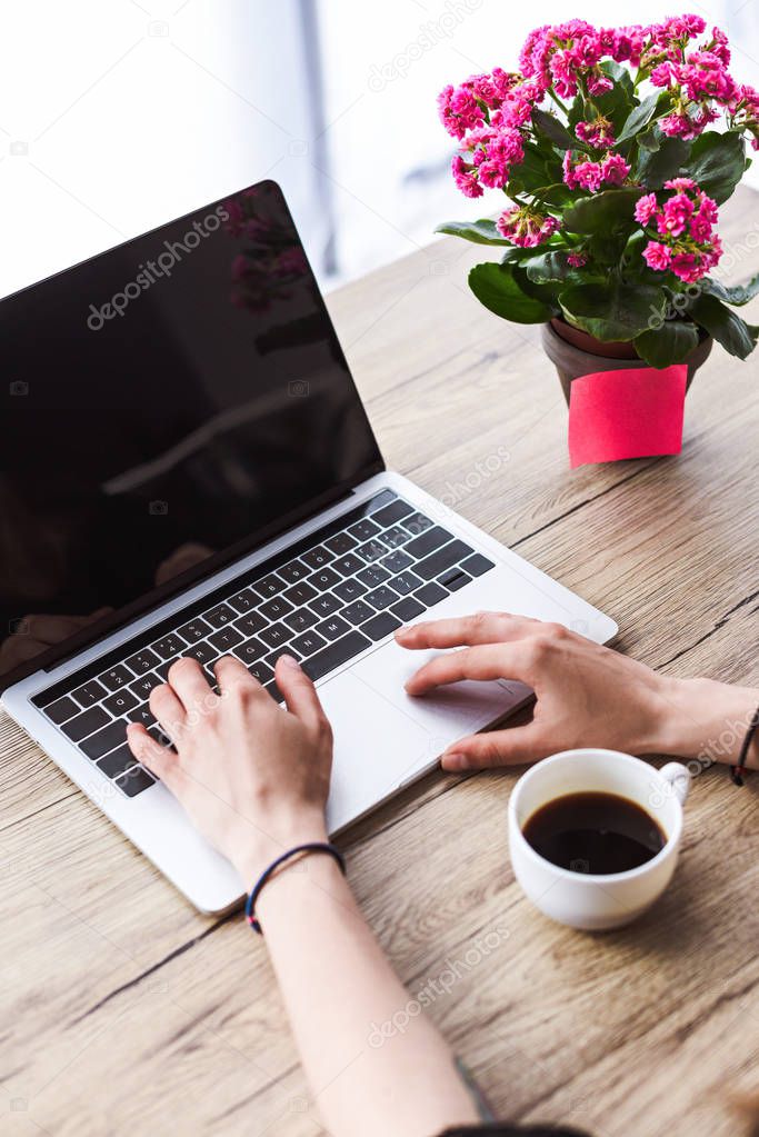 cropped shot of woman typing on laptop with blank screen at table with coffee cup and flowers 