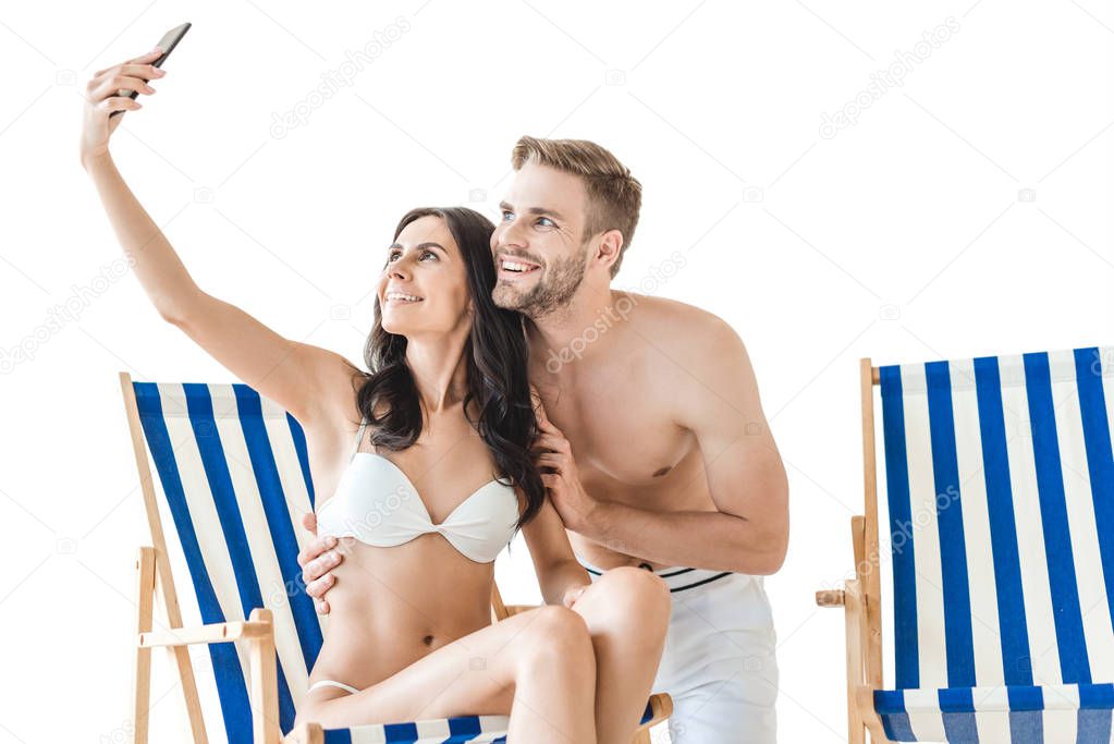 smiling couple taking selfie with smartphone on beach chairs, isolated on white