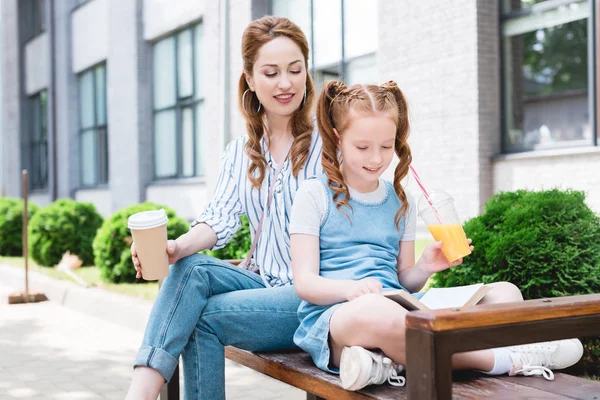 子供を浮かべてジュース路上一緒にベンチで休んでいる間母のそばで本を読んで — ストック写真