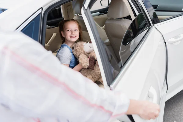 有选择的焦点微笑的孩子与泰迪熊看母亲打开车门 — 免费的图库照片