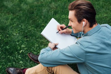 high angle view of smiling young man in earphones taking notes in blank notebook and looking away while sitting on grass clipart