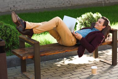 handsome smiling young freelancer lying on bench and using laptop clipart