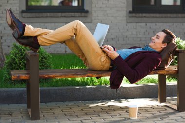 smiling young freelancer lying on bench and using laptop clipart