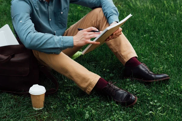 坐在草地上的年轻男子在笔记本上写的裁剪镜头 — 图库照片
