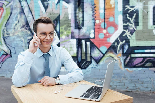 Guapo Sonriente Joven Freelancer Hablando Por Teléfono Inteligente Utilizando Ordenador —  Fotos de Stock