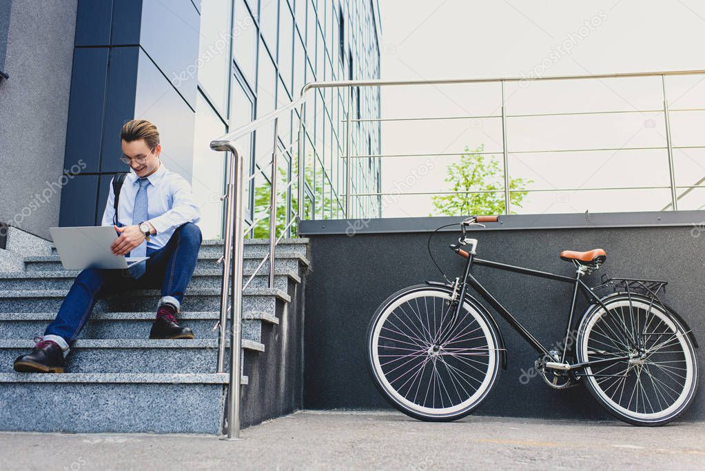 low angle view of smiling young freelancer in eyeglasses sitting on stairs and using laptop  