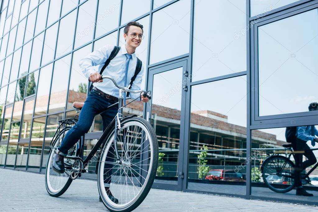 handsome smiling young man in eyeglasses and formal wear riding bike on street