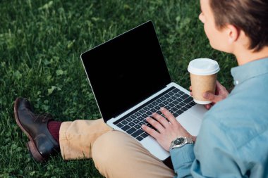 cropped shot of freelancer drinking coffee and using laptop on grass clipart