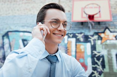 smiling young businessman in eyeglasses talking by smartphne and looking away on street clipart