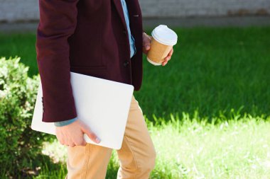 cropped shot of man with laptop and paper cup walking outside clipart