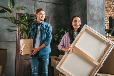 happy young woman carrying canvas and her boyfriend holding ficus plant while moving into new home clipart