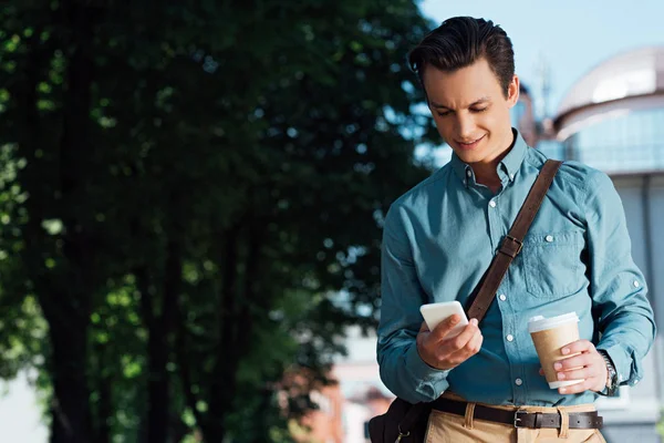 Красивый Улыбающийся Молодой Человек Держит Бумажную Чашку Использует Смартфон — стоковое фото