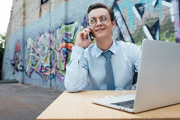 smiling young man in eyeglasses talking by smartphone and using laptop