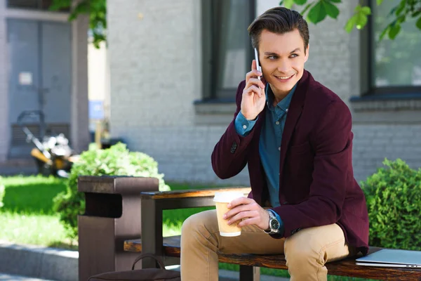 Schöner Lächelnder Junger Mann Mit Pappbecher Und Smartphone Sitzen — kostenloses Stockfoto