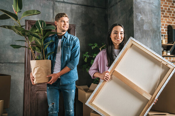 happy young woman carrying canvas and her boyfriend holding ficus plant while moving into new home