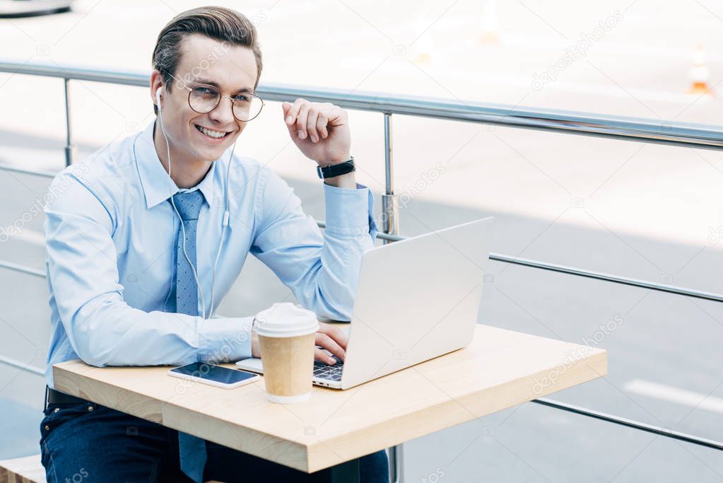 smiling young businessman in eyeglasses using laptop and earphones outside