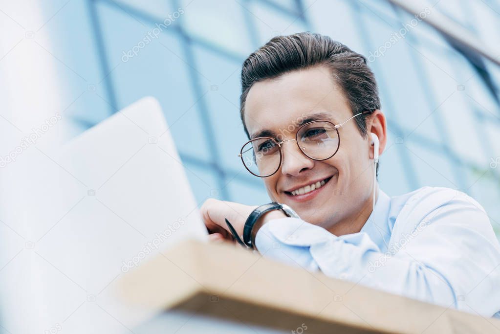 handsome smiling businessman in eyeglasses using laptop outside, selective focus
