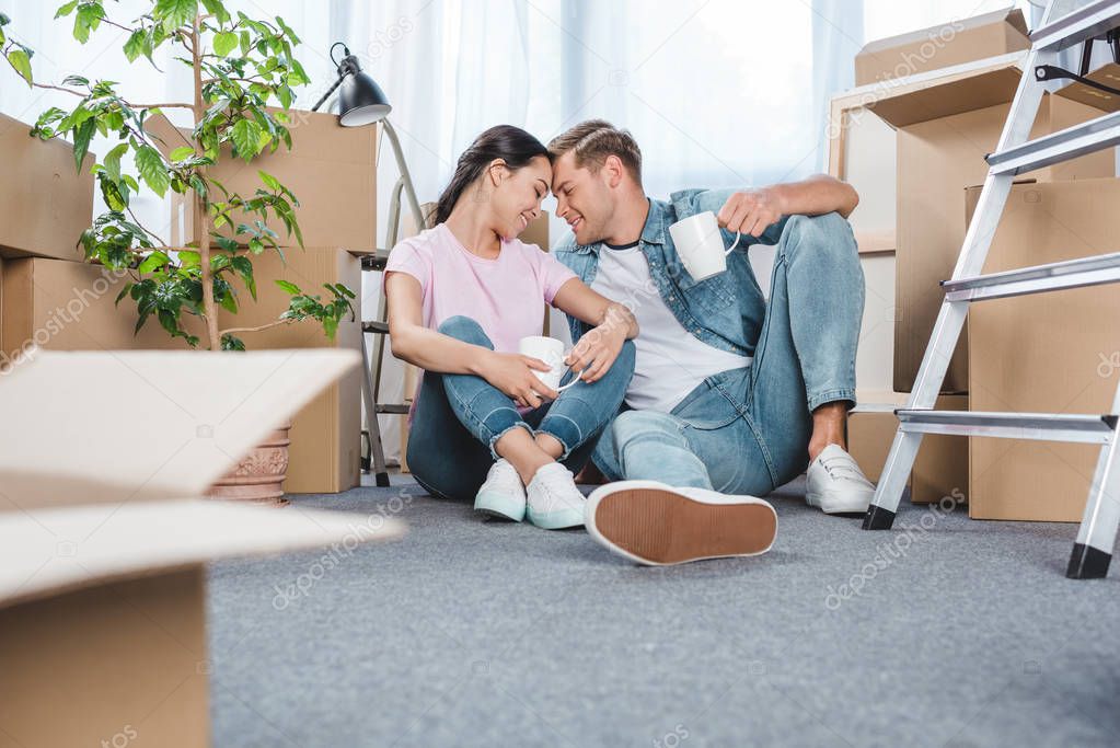 beautiful young couple with mugs sitting on floor at new home