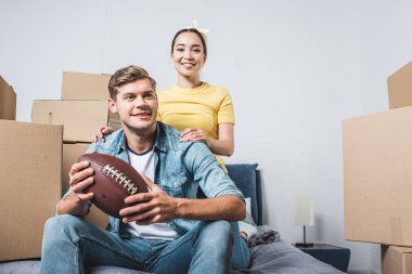 Yeni evine taşıdıktan sonra Amerikan futbolu topu ile yatakta oturmuş Interracial genç çift