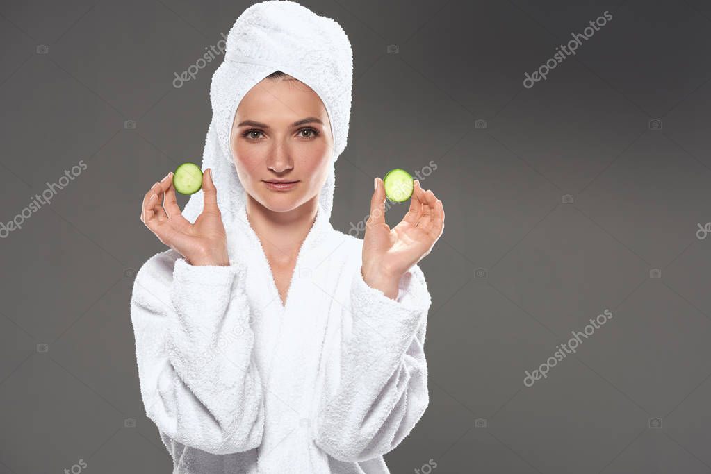 beautiful girl with cucumber slices in white bathrobe and towel, isolated on grey