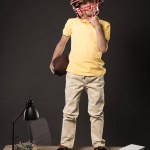 Scolaretto in casco da calcio americano con palla in mano e in piedi su tavolo con libri, piante, lampade, matite a colori, mela, orologio e libro di testo su sfondo grigio