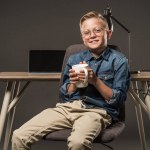 Smiling little boy in eyeglasses sitting on chair with coffee cup near table with laptop and lamp on grey background