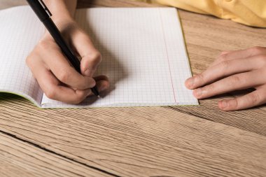 cropped image of schoolboy doing homework in empty textbook at table clipart