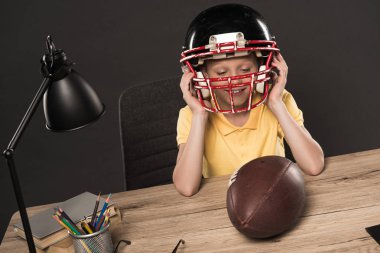 smiling schoolboy putting on american football helmet at table with ball, lamp, colour pencils and books on grey background  clipart