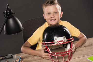 Amerikan futbol kaskı gözlük, lamba, renkli kalemler ve gri arka plan üzerinde kitap ile masada oturuyor mutlu okul çocuğu 