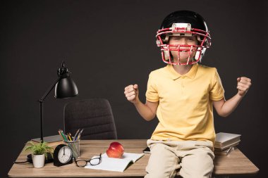 Gözlük, kitap, bitki, lamba, renkli kalem, elma, saat ve gri arka plan üzerine ders kitabı ile masada oturan Amerikan futbol kask içinde gülümseyen okul çocuğu 