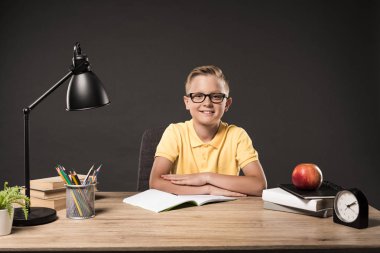 smiling schoolboy in eyeglasses with folded arms sitting at table with books, plant, lamp, colour pencils, apple, clock and textbook on grey background  clipart