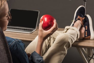 seçici odak elma tutarak ve ayaklı masada dizüstü bilgisayar ve lamba gri arka plan üzerinde oturan gözlük küçük çocuk 