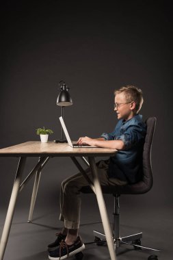 side view of happy schoolboy using laptop at table with lamp and potted plant on grey background  clipart