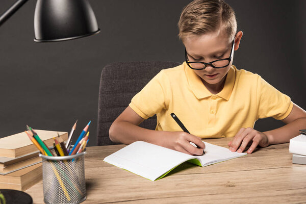 focused schoolboy doing homework in textbook at table with colour pencils, lamp and stack of books on grey background 