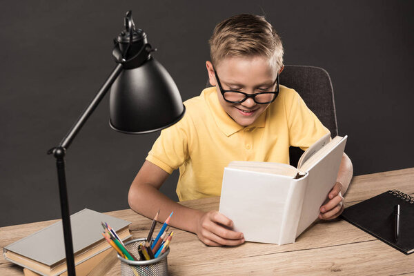 schoolboy in eyeglasses reading book at table with colour pencils, books, textbook and lamp on grey background 