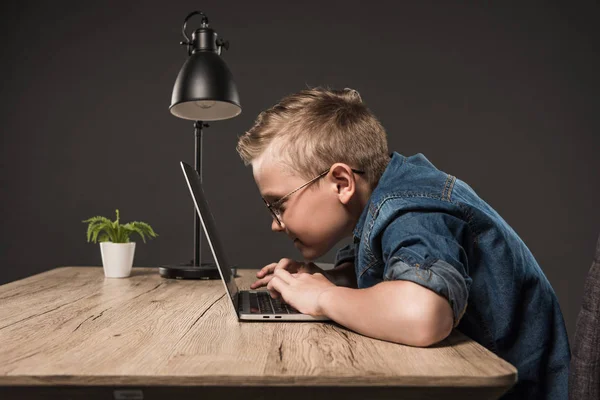 ランプと灰色の背景に植物のテーブルでノート パソコンを用いて眼鏡の男の子の側面図 — ストック写真