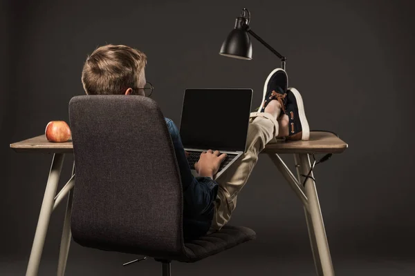 灰色の背景の空白の画面を持つノート パソコンを使用して テーブルの脚を持った少年の背面図 — ストック写真