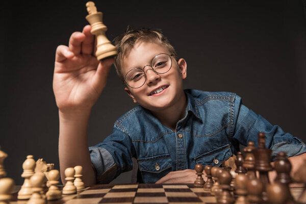 Улыбающийся мальчик в очках, держащий шахматную фигуру над шахматной доской, изолированной на сером фоне
 