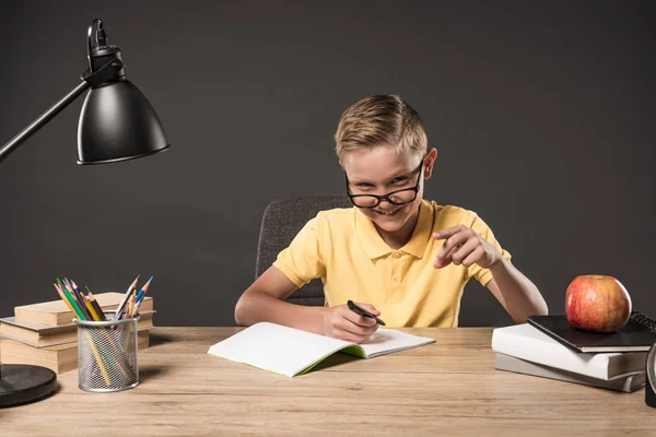 快乐的小学生在眼镜指着手指和做家庭作业在桌子上 彩色铅笔和教科书的灰色背景 — 图库照片