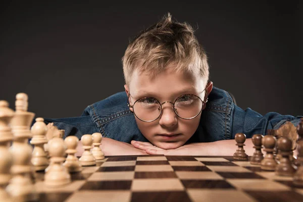 serious little boy in eyeglasses looking at camera and sitting at table with chess board isolated on grey background
