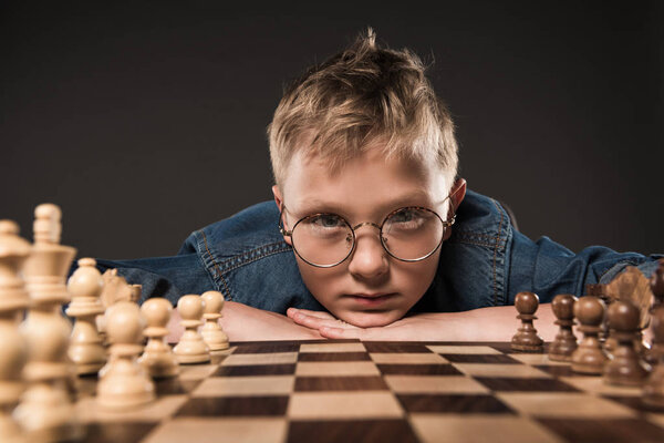 serious little boy in eyeglasses looking at camera and sitting at table with chess board isolated on grey background 