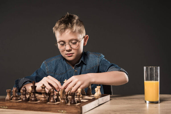 focused little boy playing chess at table with glass of juice on grey background 