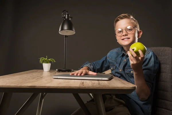 快乐的小男孩在眼镜拿着梨在桌与膝上型电脑 植物和灯在灰色背景下 — 图库照片